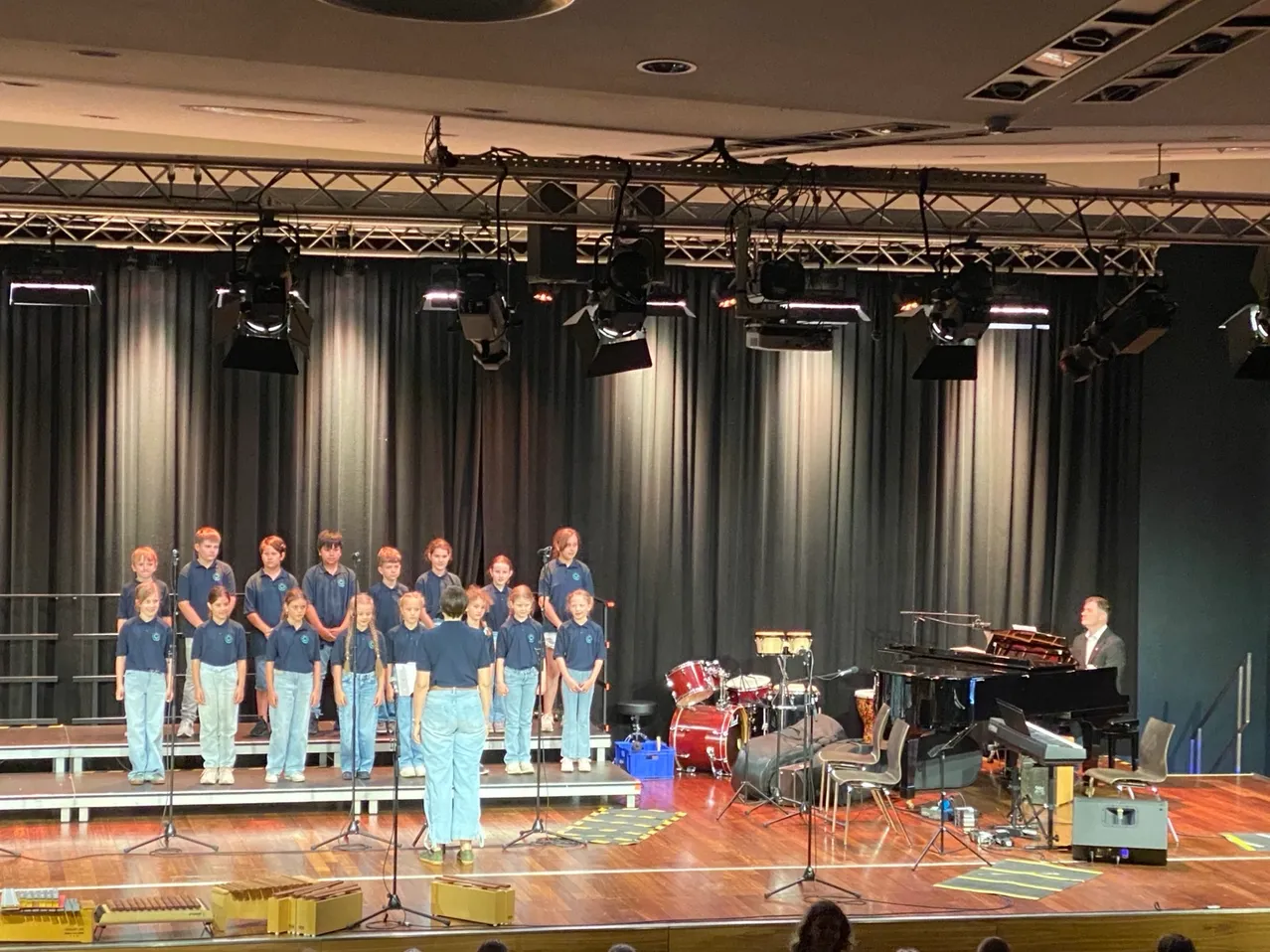 Ein Chor in blauen Uniformen tritt auf der Bühne auf, mit einem Pianisten am Flügel, Trommeln und Perkussionsinstrumenten in der Nähe. Scheinwerfer hängen von der Decke.