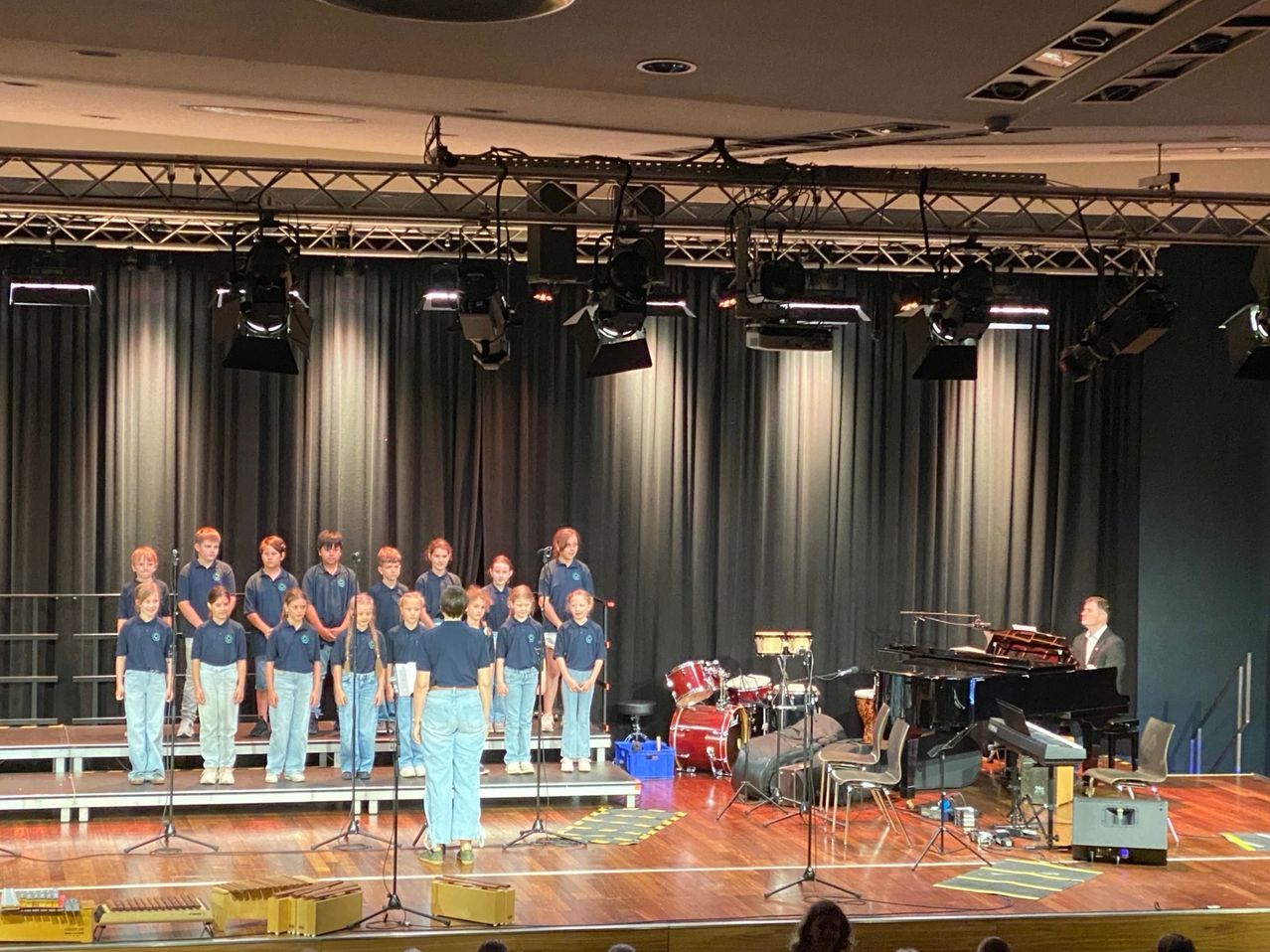 Ein Chor in blauen Uniformen tritt auf der Bühne auf, mit einem Pianisten am Flügel, Trommeln und Perkussionsinstrumenten in der Nähe. Scheinwerfer hängen von der Decke.