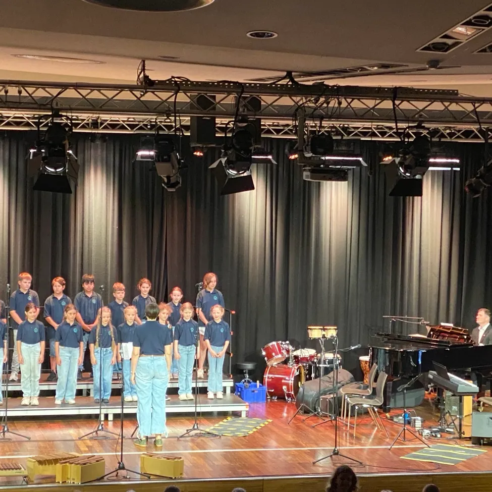 Ein Chor in blauen Uniformen tritt auf der Bühne auf, mit einem Pianisten am Flügel, Trommeln und Perkussionsinstrumenten in der Nähe. Scheinwerfer hängen von der Decke.