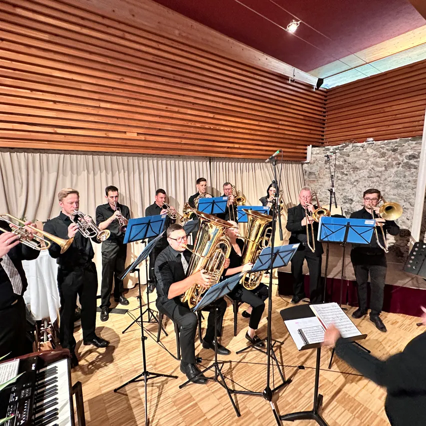 Eine Brassband steht auf der Bühne, mit Musikern, die Trompeten, Posaunen und Tuben spielen. Sie haben Notenständer und einen Pianisten zu ihrer Linken. Die Bühne ist aus Holz mit einer Steinwand im Hintergrund.