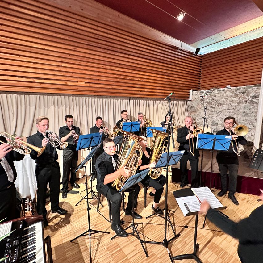Eine Brassband steht auf der Bühne, mit Musikern, die Trompeten, Posaunen und Tuben spielen. Sie haben Notenständer und einen Pianisten zu ihrer Linken. Die Bühne ist aus Holz mit einer Steinwand im Hintergrund.