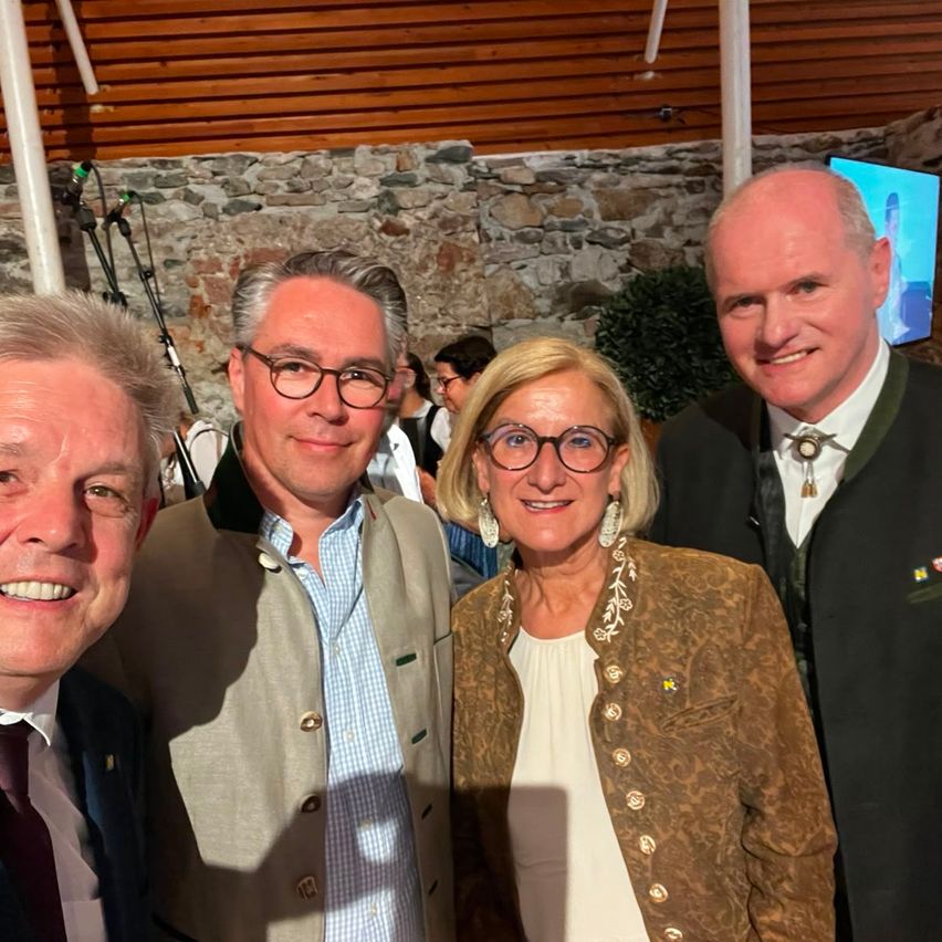 Vier Personen, zwei Männer und zwei Frauen, lächeln für ein Foto. Der Mann links trägt eine Krawatte. Die Frau in der Mitte hat eine gemusterte Jacke. Die anderen beiden Männer tragen Anzüge.