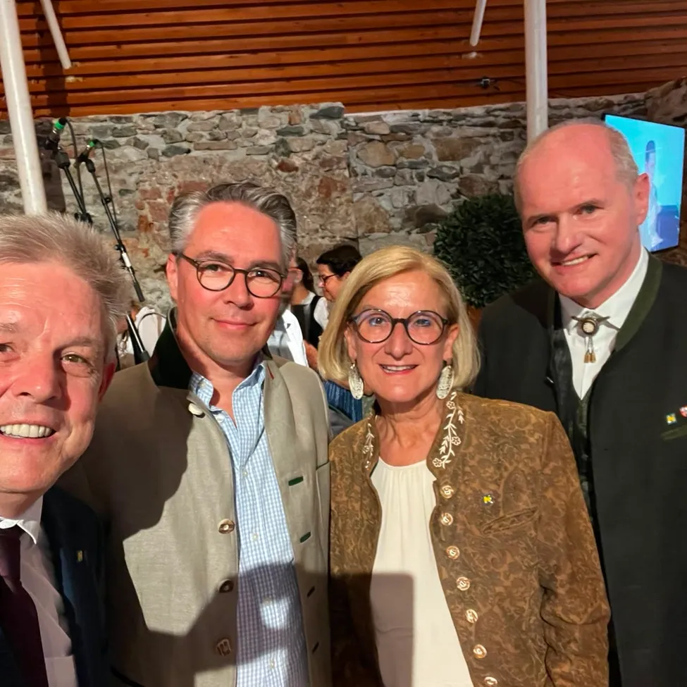 Vier Personen, zwei Männer und zwei Frauen, lächeln für ein Foto. Der Mann links trägt eine Krawatte. Die Frau in der Mitte hat eine gemusterte Jacke. Die anderen beiden Männer tragen Anzüge.