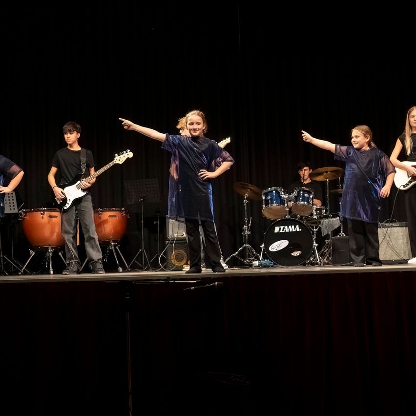 Eine Band tritt auf der Bühne mit Trommeln, Gitarren und Becken auf. Zwei Mädchen und ein Junge spielen Gitarren, während ein anderer Junge Schlagzeug spielt.