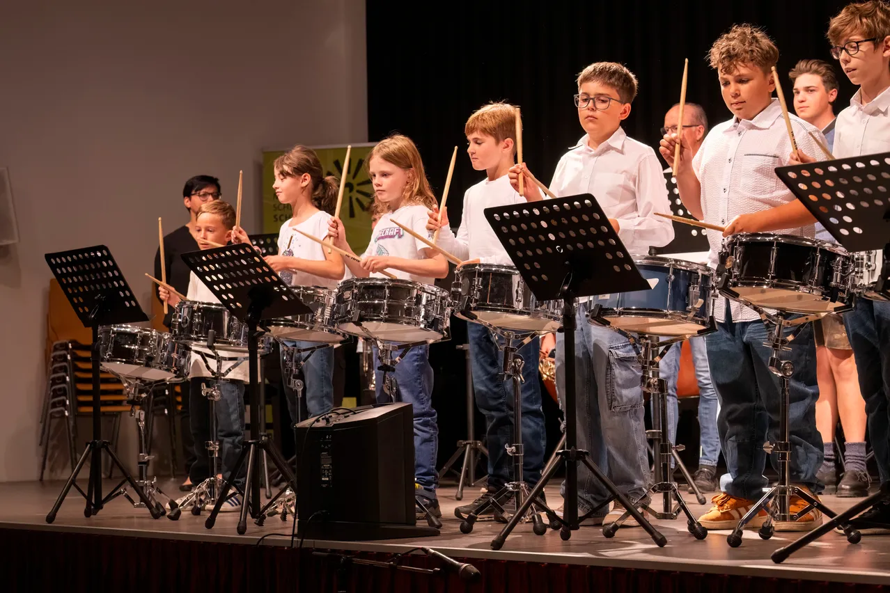 Eine Gruppe junger Kinder tritt mit Trommeln und Drumsticks auf der Bühne auf. Sie sind in weißen Hemden und Jeans gekleidet. Hinter ihnen befindet sich ein schwarzer Hintergrund und ein grünes Banner.