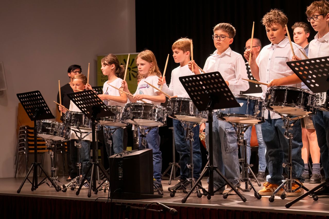 Eine Gruppe junger Kinder tritt mit Trommeln und Drumsticks auf der Bühne auf. Sie sind in weißen Hemden und Jeans gekleidet. Hinter ihnen befindet sich ein schwarzer Hintergrund und ein grünes Banner.