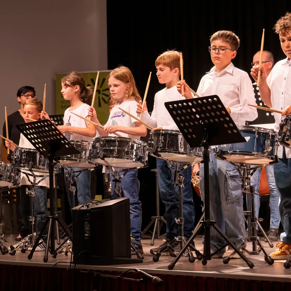 Eine Gruppe junger Kinder tritt mit Trommeln und Drumsticks auf der Bühne auf. Sie sind in weißen Hemden und Jeans gekleidet. Hinter ihnen befindet sich ein schwarzer Hintergrund und ein grünes Banner.