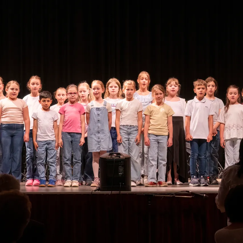 Eine Gruppe von Kindern steht auf der Bühne und ist wahrscheinlich bei einer Schulveranstaltung. Sie tragen Freizeitkleidung und einige von ihnen tragen eine Brille. Ein Lautsprecher steht vor ihnen.