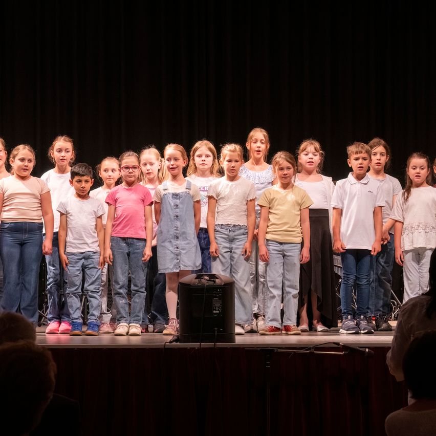Eine Gruppe von Kindern steht auf der Bühne und ist wahrscheinlich bei einer Schulveranstaltung. Sie tragen Freizeitkleidung und einige von ihnen tragen eine Brille. Ein Lautsprecher steht vor ihnen.
