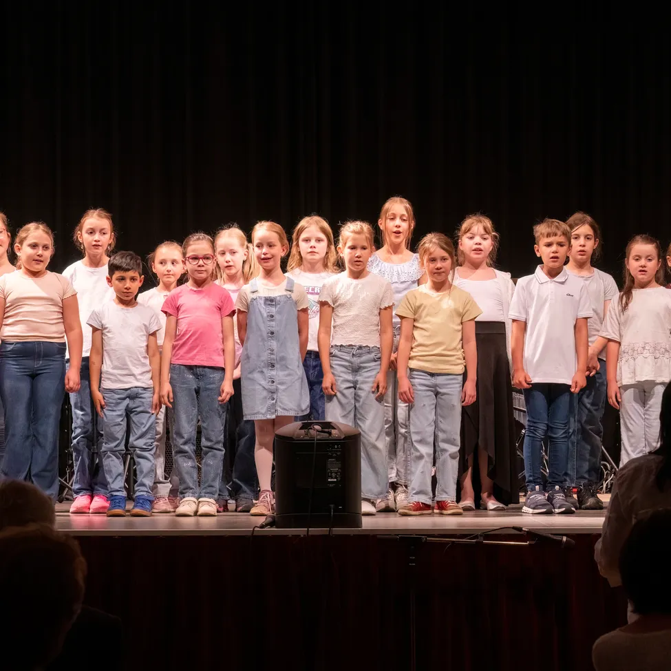 Eine Gruppe von Kindern steht auf der Bühne und ist wahrscheinlich bei einer Schulveranstaltung. Sie tragen Freizeitkleidung und einige von ihnen tragen eine Brille. Ein Lautsprecher steht vor ihnen.