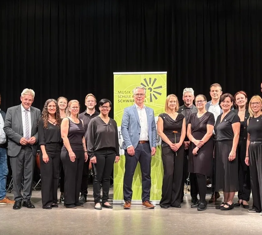Gruppenfoto von Erwachsenen in schwarzer Kleidung, die auf der Bühne für ein Foto posieren, mit einem grünen Banner mit der Aufschrift 'Musik Schulen Schwarz'.
