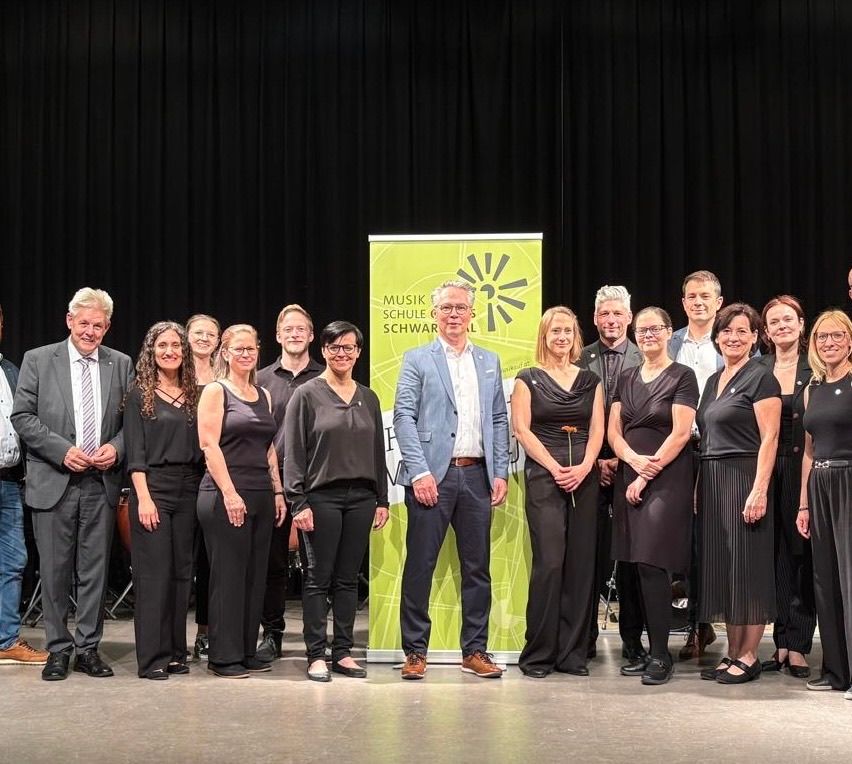 Gruppenfoto von Erwachsenen in schwarzer Kleidung, die auf der Bühne für ein Foto posieren, mit einem grünen Banner mit der Aufschrift 'Musik Schulen Schwarz'.