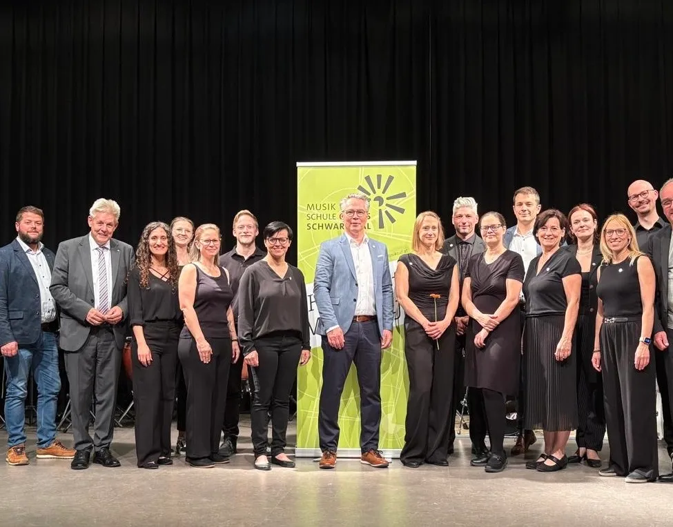 Gruppenfoto von Erwachsenen in schwarzer Kleidung, die auf der Bühne für ein Foto posieren, mit einem grünen Banner mit der Aufschrift 'Musik Schulen Schwarz'.