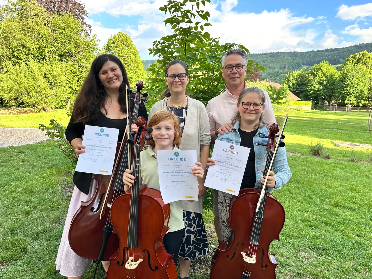 Eine Familie mit Kindern, alle Cellos und Urkunden in der Hand, steht auf einem Rasenfeld unter einem Baum. Die Kinder lächeln und die Erwachsenen schauen in die Kamera.