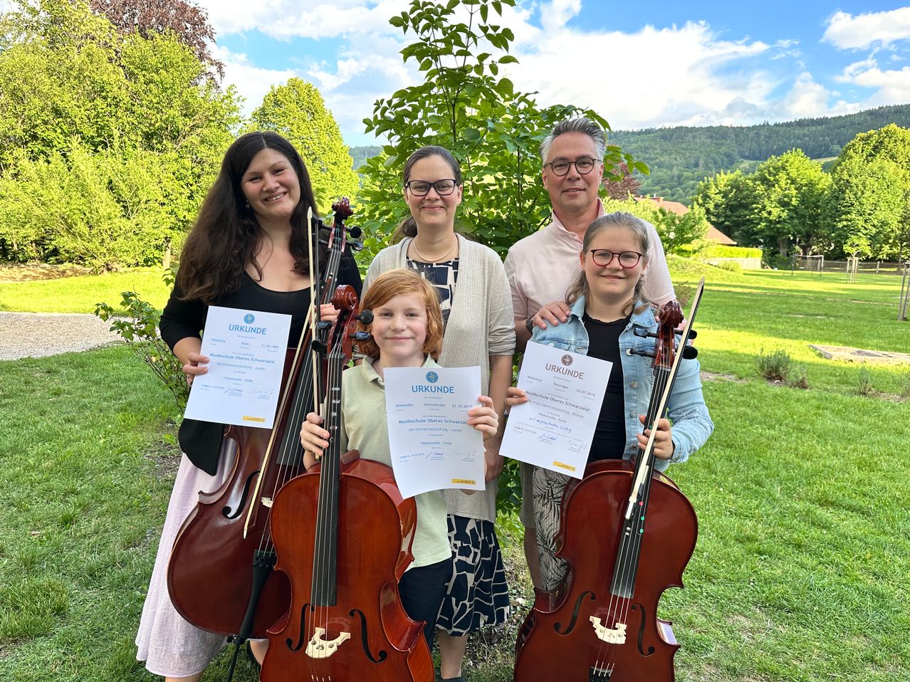 Eine Familie mit Kindern, alle Cellos und Urkunden in der Hand, steht auf einem Rasenfeld unter einem Baum. Die Kinder lächeln und die Erwachsenen schauen in die Kamera.