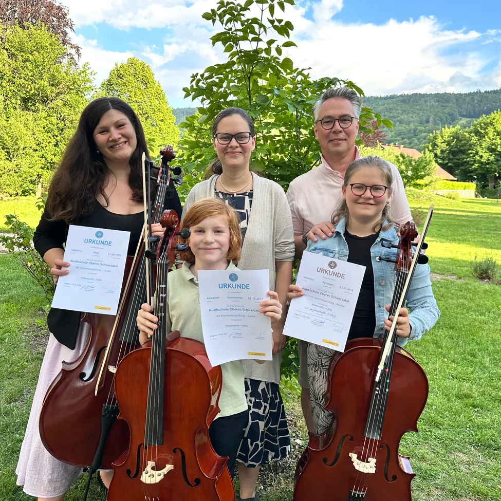 Eine Familie mit Kindern, alle Cellos und Urkunden in der Hand, steht auf einem Rasenfeld unter einem Baum. Die Kinder lächeln und die Erwachsenen schauen in die Kamera.