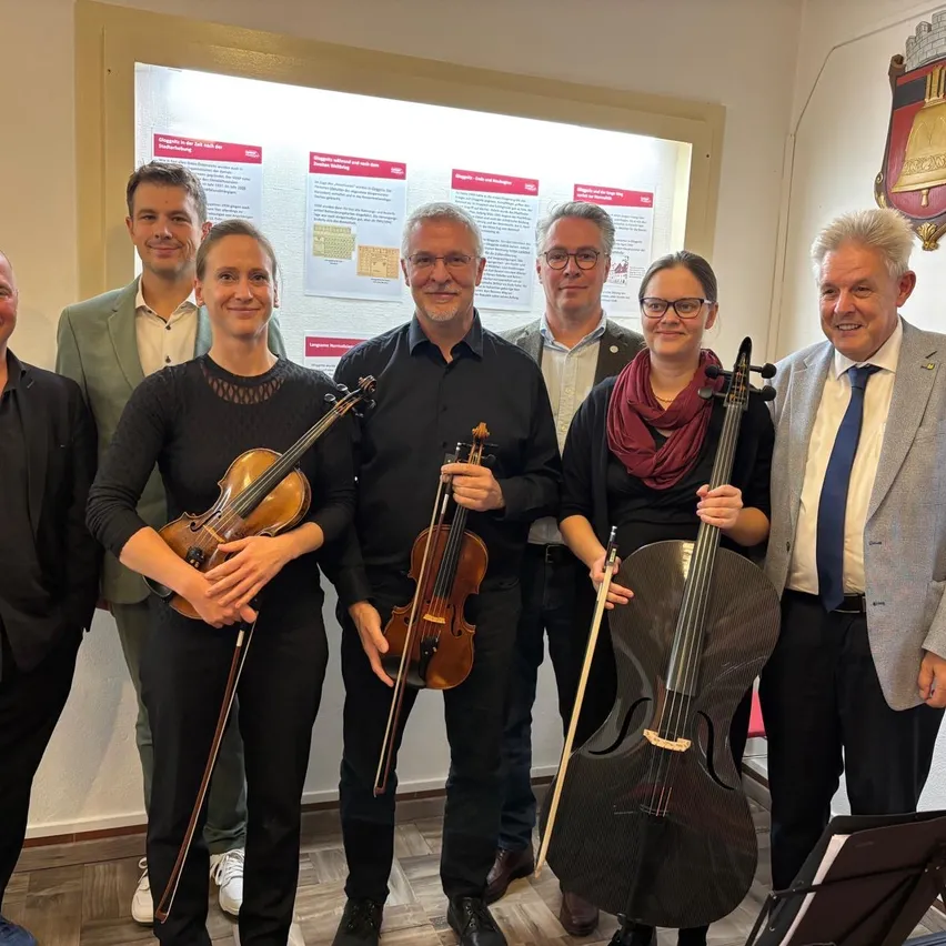Eine Gruppe von Menschen posiert für ein Foto mit Violinen und einem Cello. Sie stehen vor einer weißen Wand mit einem Poster und einem Wappen.