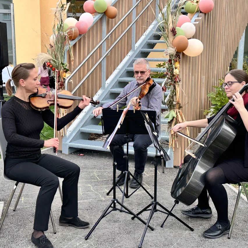 Drei Musiker spielen auf einer Bühne. Eine Frau spielt die Geige, ein Mann spielt die Geige und eine andere Frau spielt das Cello. Sie sitzen auf Stühlen vor einer Treppe mit bunten Luftballons.