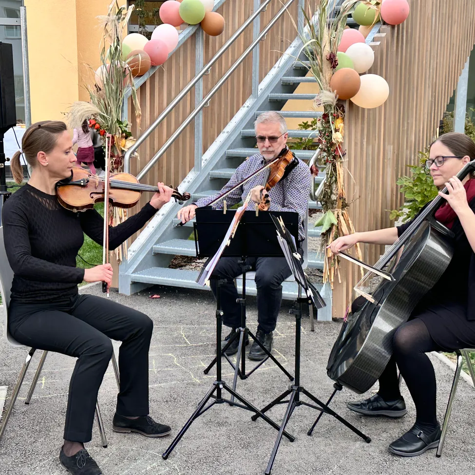 Drei Musiker spielen auf einer Bühne. Eine Frau spielt die Geige, ein Mann spielt die Geige und eine andere Frau spielt das Cello. Sie sitzen auf Stühlen vor einer Treppe mit bunten Luftballons.
