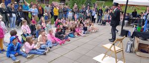 Eine Gruppe von Menschen, einschließlich Kindern, sitzt auf dem Boden, während ein Mann vor ihnen steht und wahrscheinlich eine Rede hält. Sie befinden sich in einem Außenbereich mit Betonboden.