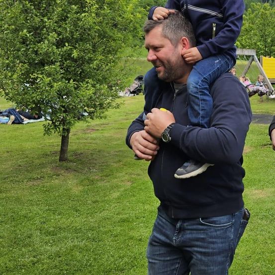 Ein Mann trägt ein Kind auf seinen Schultern im Park und lächelt. In der Nähe steht ein Kinderwagen, und einige Kinder spielen auf einer Schaukel.