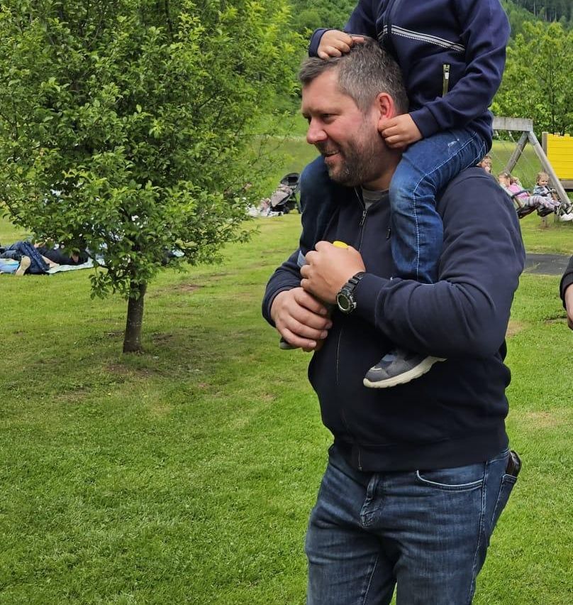 Ein Mann trägt ein Kind auf seinen Schultern im Park und lächelt. In der Nähe steht ein Kinderwagen, und einige Kinder spielen auf einer Schaukel.