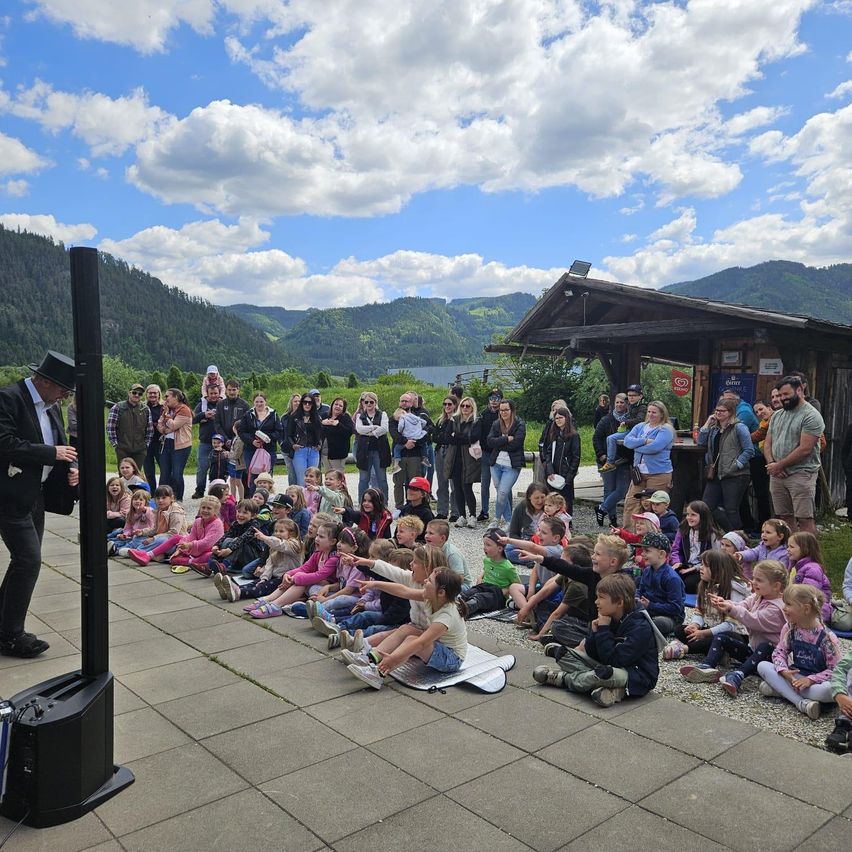 Ein Mann in Anzug steht auf einer Bühne und spricht eine Menschenmenge an, die auf dem Boden sitzt. Dahinter befindet sich ein See und Berge unter einem bewölkten Himmel.