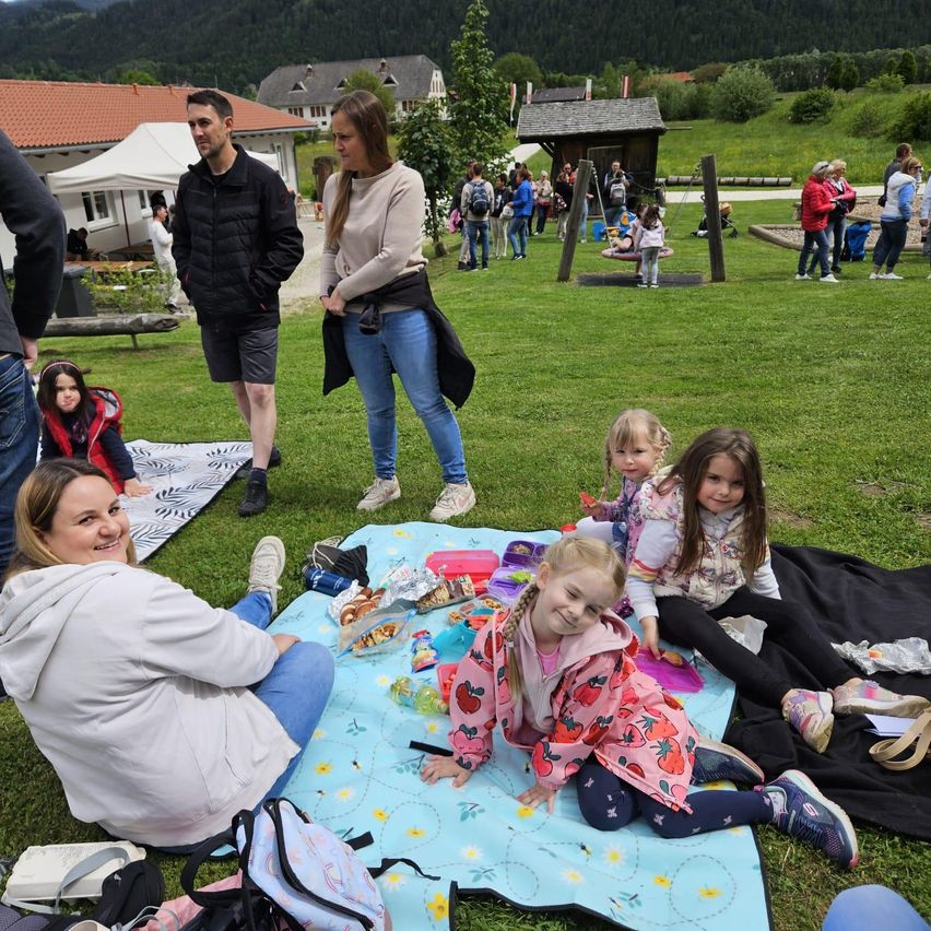 Eine Familie mit Kindern macht ein Picknick auf einem Rasen mit Bergkulisse. Einige Leute stehen und unterhalten sich. In der Nähe spielen andere mit Kindern auf einem Spielplatz.
