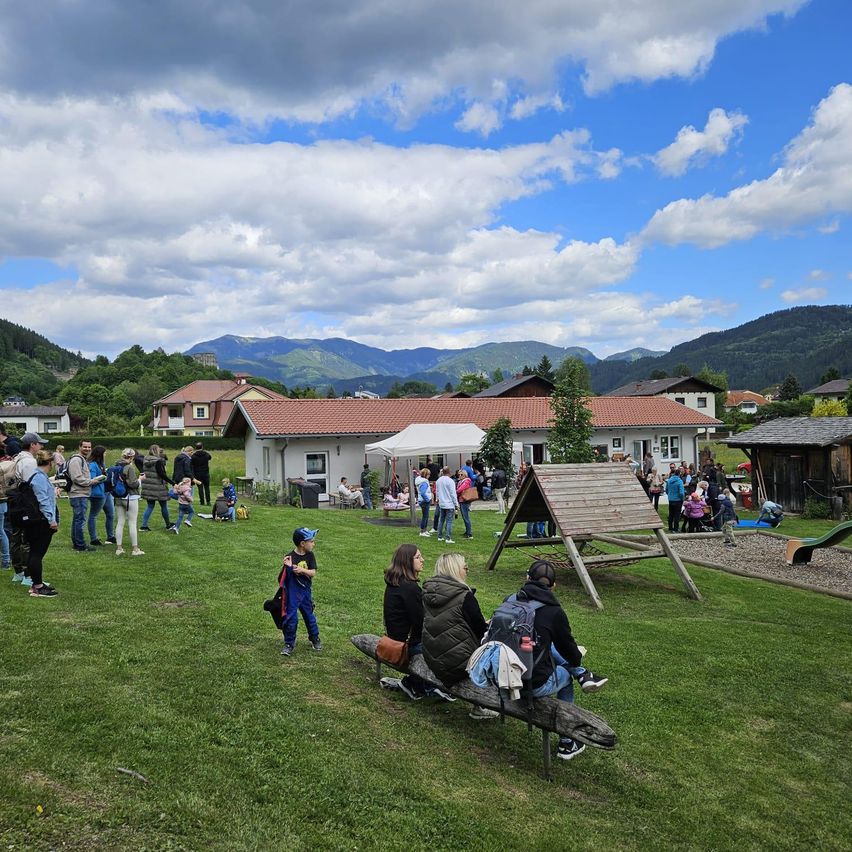 Eine Gruppe von Menschen versammelt sich auf einer Rasenfläche, einige sitzen auf einer Bank, andere gehen oder stehen. Ein Haus mit rotem Dach im Hintergrund, vor dem ein Zelt steht. In der Ferne befinden sich Berge und Bäume.