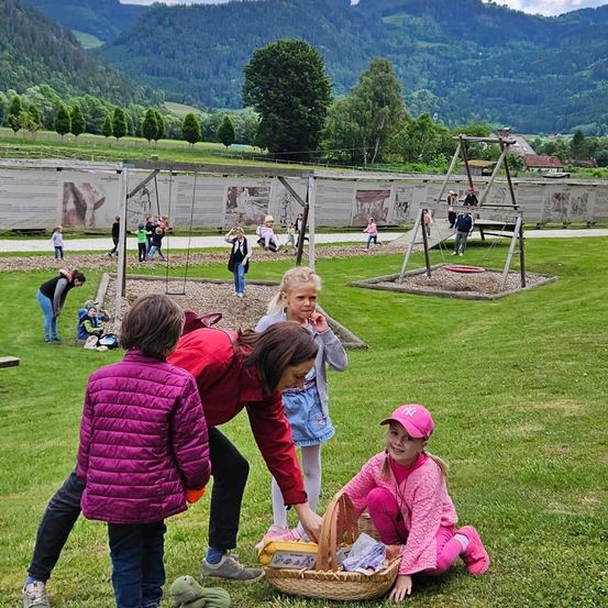Eine Gruppe von Kindern spielt in einem Park mit Bergen im Hintergrund. Einige sitzen auf dem Rasen, während andere auf den Schaukeln spielen.
