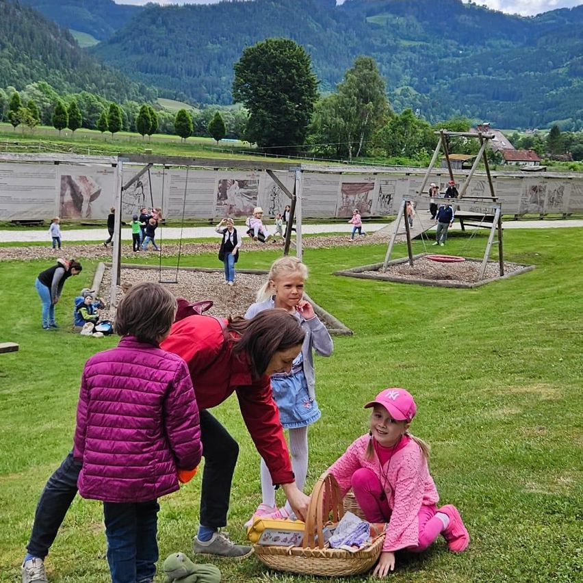 Eine Gruppe von Kindern spielt in einem Park mit Bergen im Hintergrund. Einige sitzen auf dem Rasen, während andere auf den Schaukeln spielen.