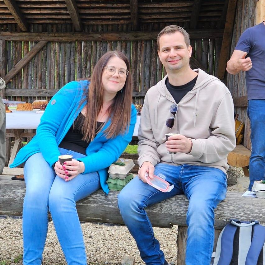 Ein Mann und eine Frau sitzen lächelnd auf einer Holzbank. Die Frau trägt eine blaue Jacke und eine Brille, während der Mann eine Kapuzenjacke trägt und einen Plastikbehälter hält. Hinter ihnen steht eine Holzstruktur mit einem Tisch.