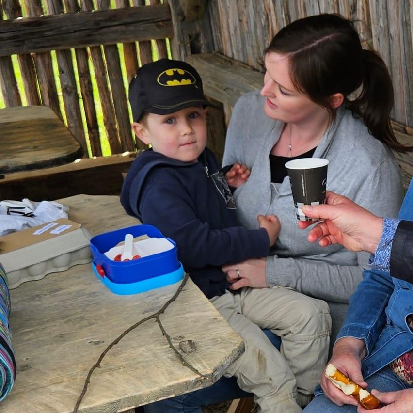 Eine Familie sitzt an einem Holztisch. Ein junger Junge trägt eine Batman-Kappe. Eine Frau hält eine Kaffeetasse. Eine andere Person reicht dem Jungen ein Essen.