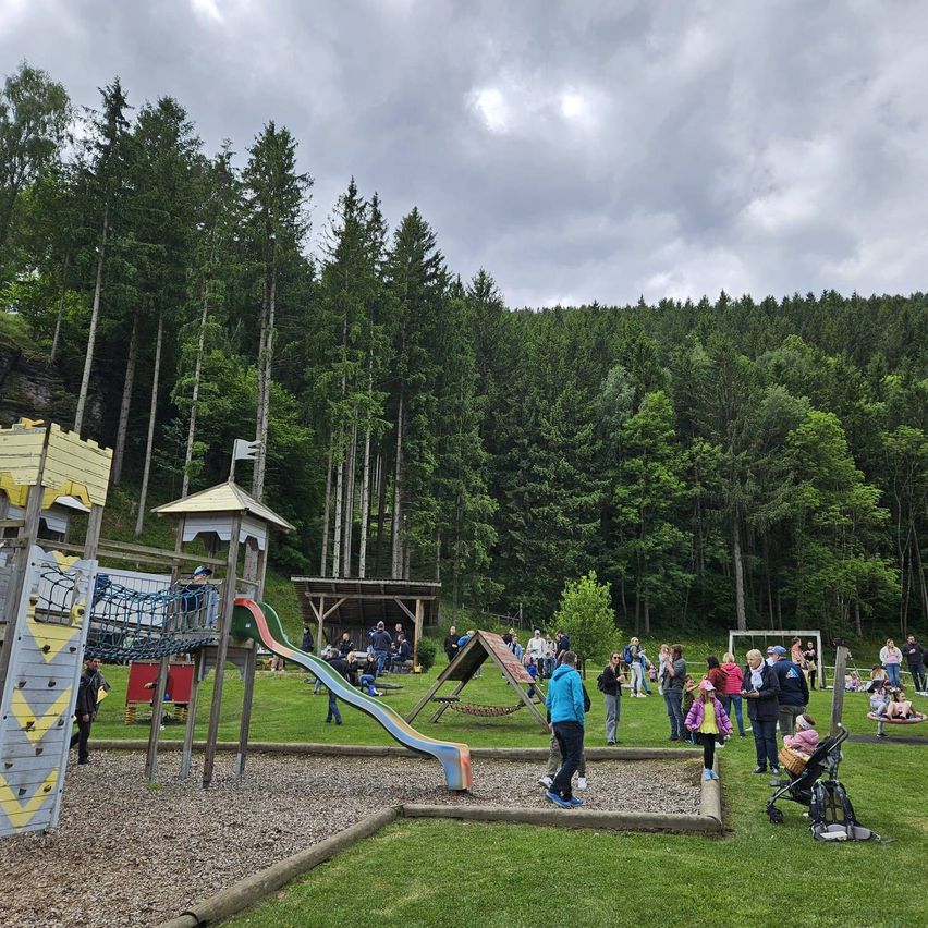 Ein Freiluftspielplatz mit Rutsche und Kletterwand ist gefüllt mit Familien. Einige Leute gehen herum, während andere auf dem Gras sitzen. Es gibt ein kleines Holzhaus und ein Fußballtor im Hintergrund.