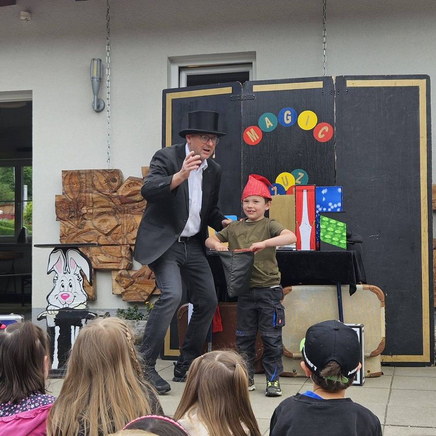 Ein Zauberer in einem Zylinder führt eine Vorstellung für eine Gruppe von Kindern durch, wobei ein Junge mit rotem Hut einen Hut hält. Dahinter steht eine Kulisse mit bunten Buchstaben.