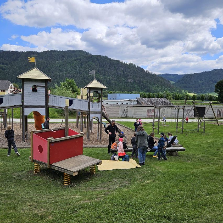 Ein Spielplatz mit einem Schloss und Rutschen. Menschen stehen und sitzen auf dem Gras. Eine Person geht. Ein Berg ist im Hintergrund.