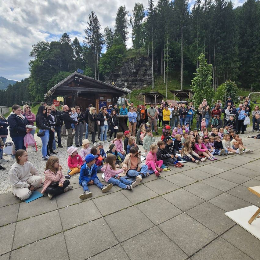 Eine Gruppe von Menschen, einschließlich Kindern, sitzt draußen auf dem Boden, mit einigen Erwachsenen in der Nähe. Im Hintergrund befindet sich ein kleines Gebäude, und Bäume und Berge sind sichtbar.