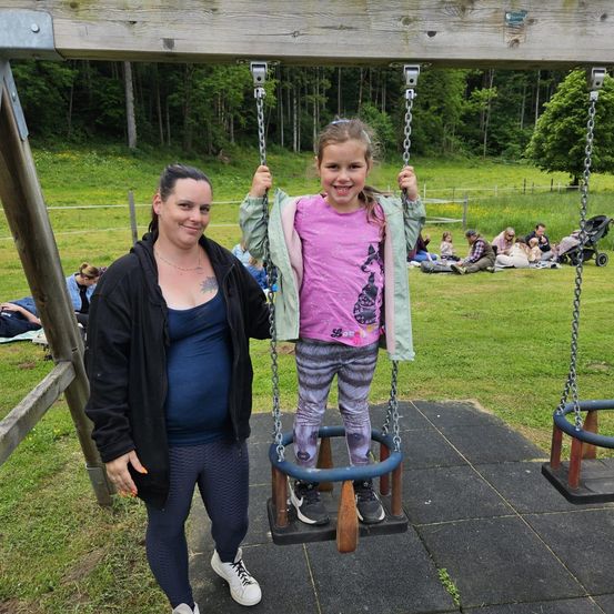 Eine schwangere Frau stützt ein kleines Mädchen auf einer Schaukel in einem Park, während andere Familien im Hintergrund die Natur genießen.