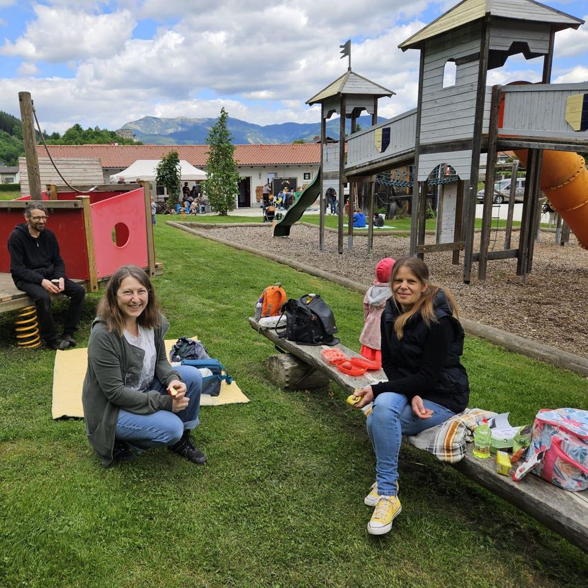 Zwei Frauen sitzen auf einer Bank im Spielplatz, während ein Mann auf einer Bank in der Nähe sitzt. Verschiedene Gegenstände, darunter Taschen und Spielzeug, sind um sie herum verstreut. Im Hintergrund spielt ein Kind auf einer Rutsche.
