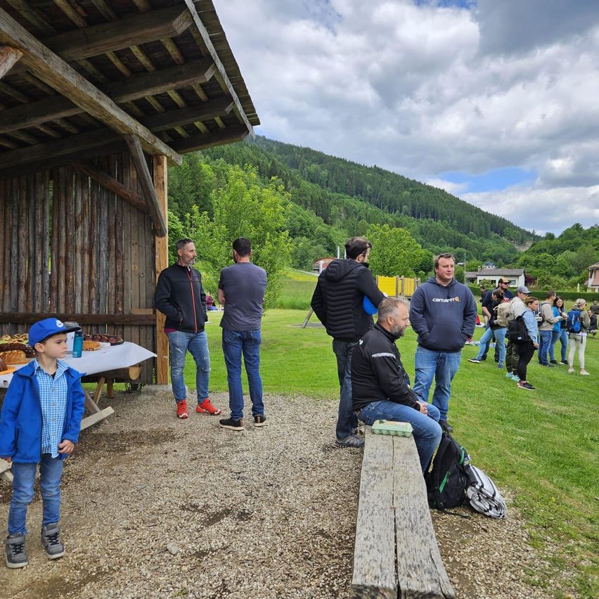 Eine Gruppe von Männern steht an einem sonnigen Tag in der Nähe einer Holzstruktur, möglicherweise einer Scheune. Ein junger Junge steht in der Nähe und schaut zu ihnen. Sie sind von einem Grasfeld umgeben, mit einem Berg im Hintergrund.