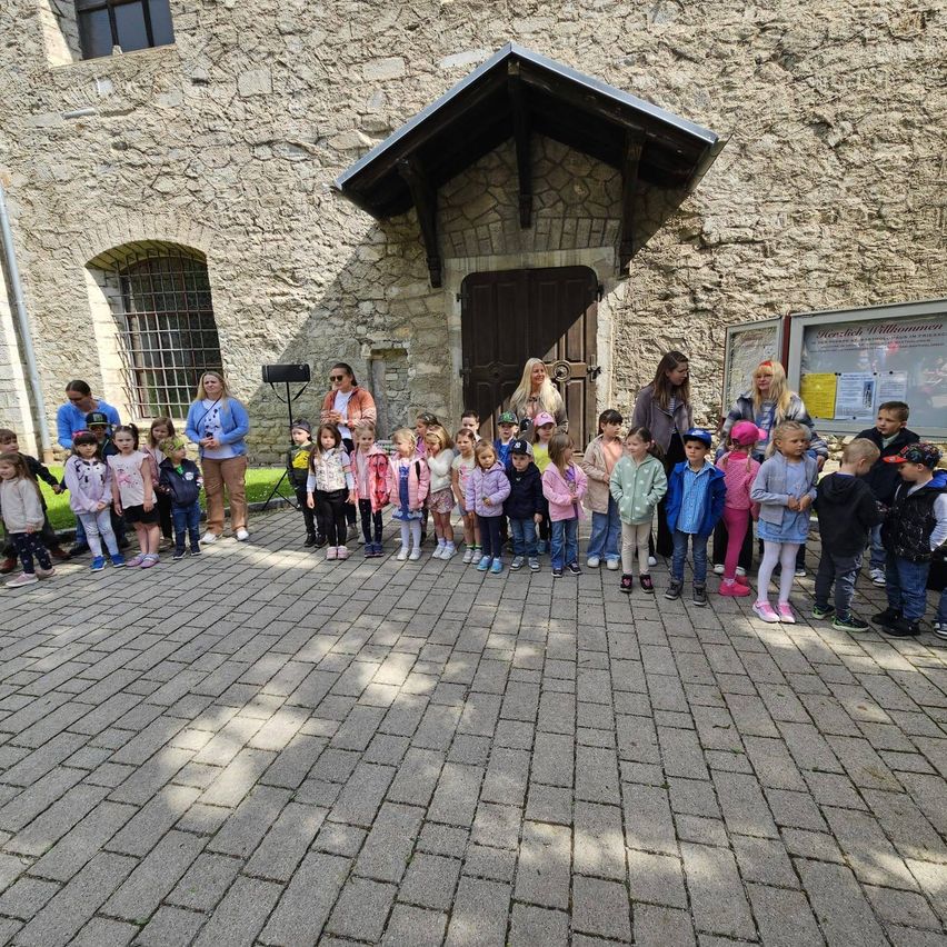 Eine Gruppe von Kindern steht in einer Reihe vor einem Steingebäude mit einem dreieckigen Dach. Einige Erwachsene sind ebenfalls anwesend, und sie tragen alle Mützen und Turnschuhe.