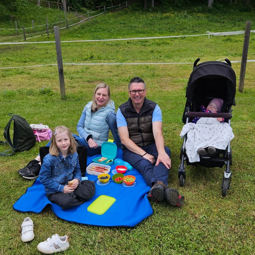 Eine Familie macht ein Picknick auf einem Grasfeld mit einem Baby im Kinderwagen, einem kleinen Mädchen, einer Frau und einem Mann, die auf einer Decke sitzen.