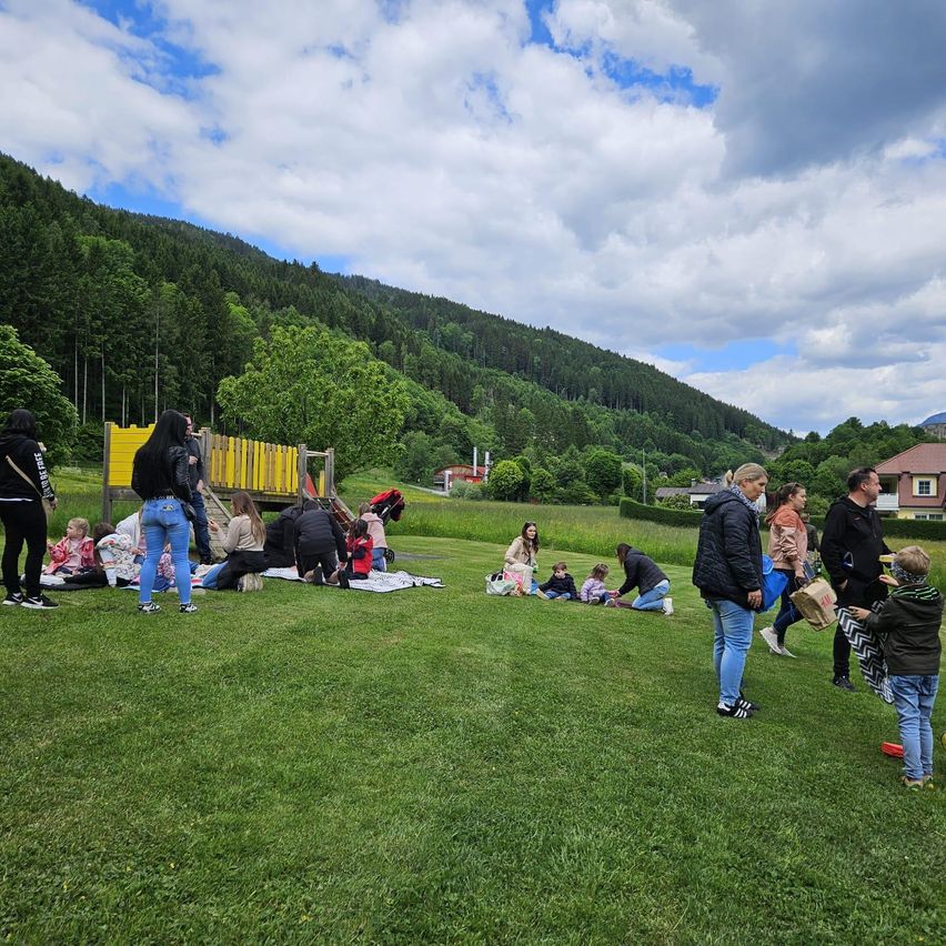 Eine Gruppe von Menschen, darunter auch Kinder, versammelt sich auf einem Rasenfeld mit einer gelben Spielstruktur und Bäumen im Hintergrund. Einige sitzen auf dem Gras, während andere stehen oder gehen. In der Ferne befinden sich Häuser und eine Bergkette.