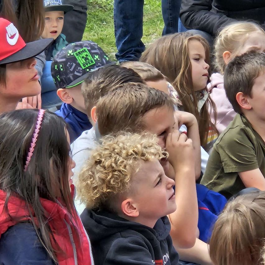 Eine Gruppe von Kindern und Erwachsenen versammelt sich im Freien, möglicherweise bei einer Sportveranstaltung. Einige Kinder tragen Mützen, während andere lange Haare haben. Ein Junge mit lockigem Haar schaut nach unten, und ein Mädchen mit einer roten Jacke beobachtet ihn.