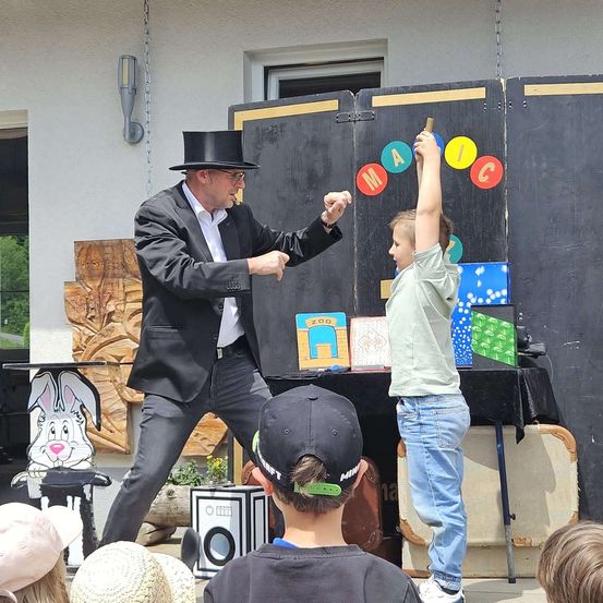 Ein Zauberer führt einen Trick vor einer Gruppe von Kindern vor. Der Zauberer trägt einen Zylinder und hält einen Zauberstab. Ein Junge hebt die Hand, während der Zauberer eine Karte aus dem Mund des Jungen zieht.