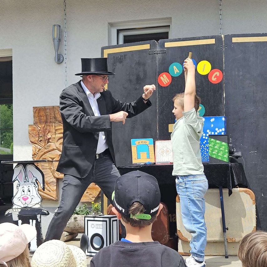 Ein Zauberer führt einen Trick vor einer Gruppe von Kindern vor. Der Zauberer trägt einen Zylinder und hält einen Zauberstab. Ein Junge hebt die Hand, während der Zauberer eine Karte aus dem Mund des Jungen zieht.