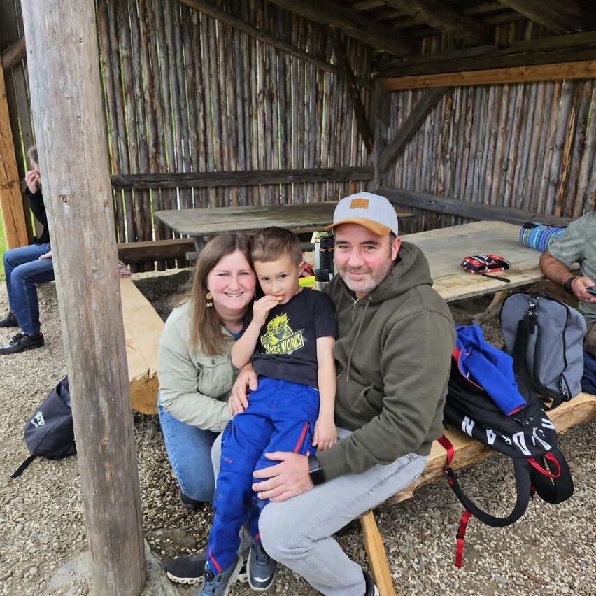 Eine Familie sitzt auf einer Holzbank in einem Unterstand. Der Mann trägt eine Kappe und eine Kapuzenjacke, und die Frau trägt eine Jacke. Der Junge hält einen Snack und lächelt. Hinter ihnen befinden sich Taschen und ein Tisch.