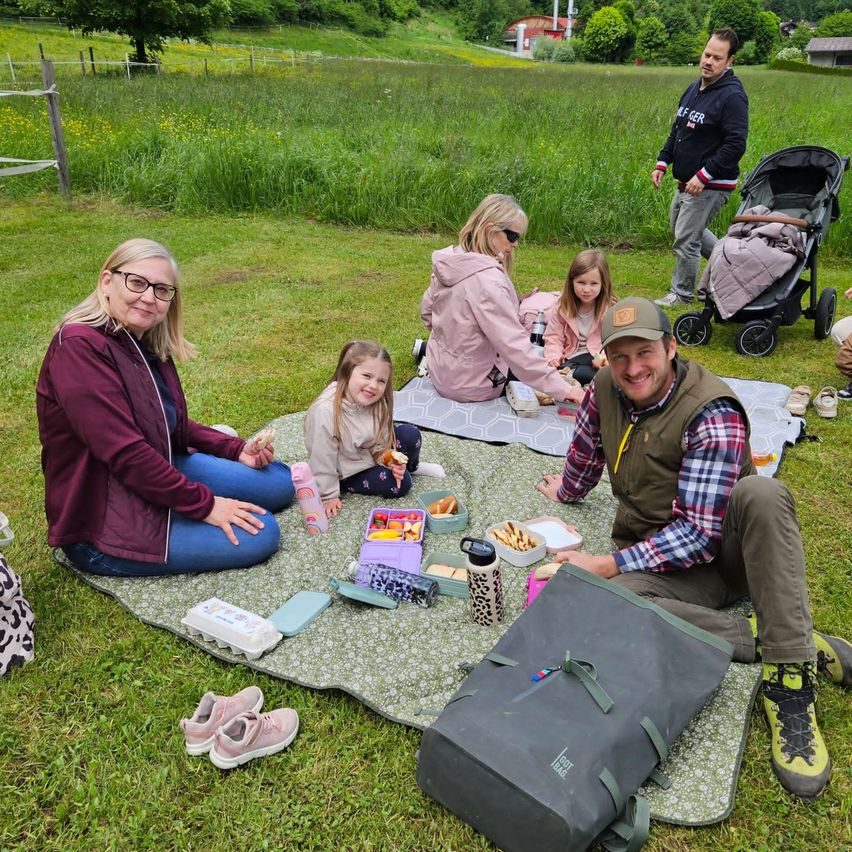 Eine Familie macht ein Picknick auf einer Wiese. Es sind mehrere Erwachsene und Kinder anwesend, zusammen mit einem Kinderwagen und einer Tasche mit Picknickutensilien.