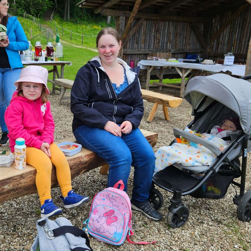 Eine Frau und ein junges Mädchen sitzen auf einer Bank in einer Outdoor-Umgebung. Das Mädchen trägt eine Mütze und Brille, und ein Baby ruht in einem Kinderwagen neben ihnen. Eine Holzstruktur und ein Tisch mit verschiedenen Gegenständen sind im Hintergrund.