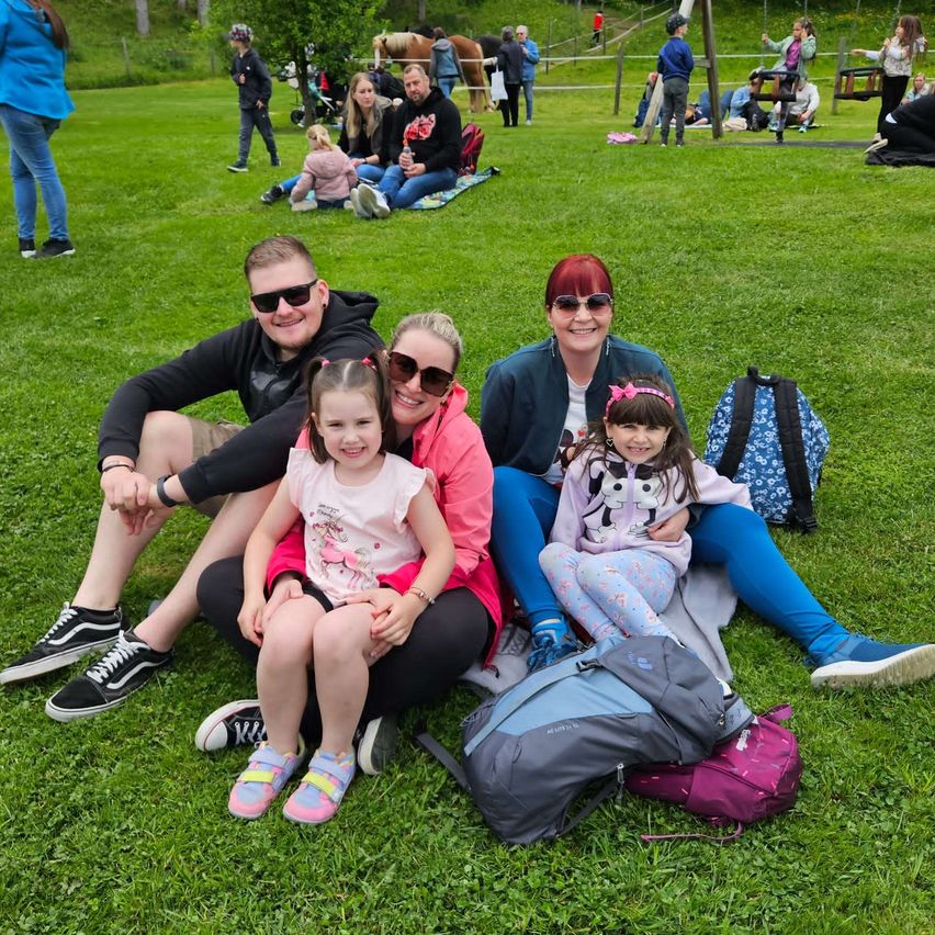 Eine Familie sitzt auf einem Rasenfeld mit zwei kleinen Mädchen, umgeben von Rucksäcken und anderen Familien, die den Park genießen.